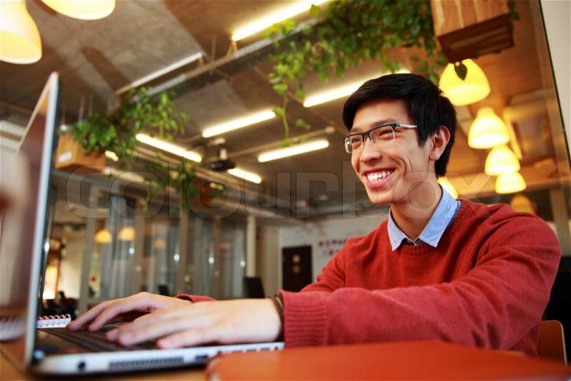 Cheerful asian man in glasses typing on laptop in office, stock photo