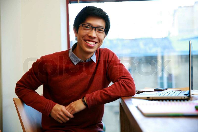 Happy asian man sitting at the table with laptop and looking at camera, stock photo