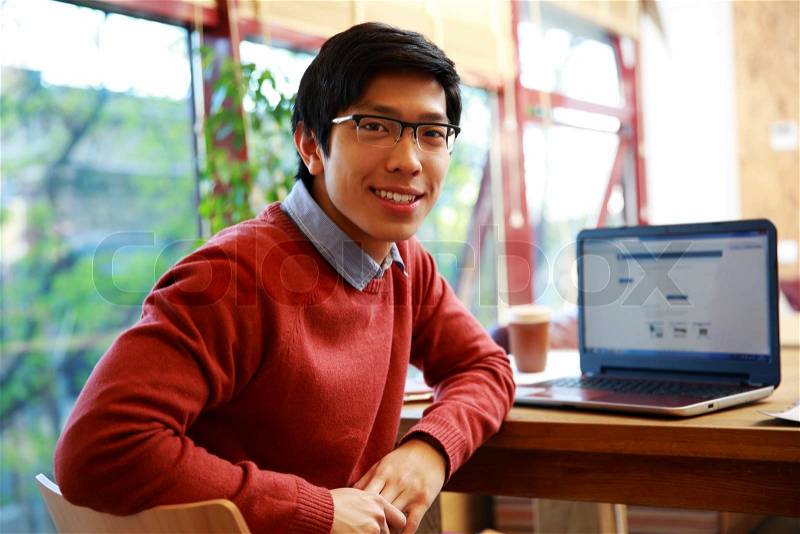 Young asian man sitting at the table with laptop and looking at camera, stock photo