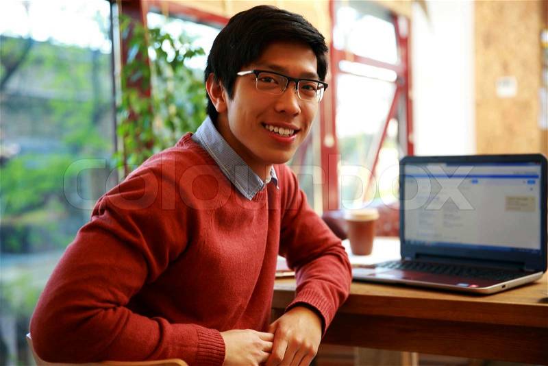 Happy asian man sitting at the table with laptop and looking at camera, stock photo