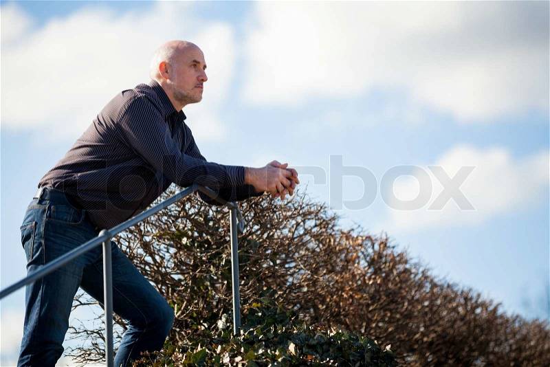 View from below of a thoughtful middle-aged man sitting on a flight of steps staring into the distance with a serious expression against a blue sky, stock photo