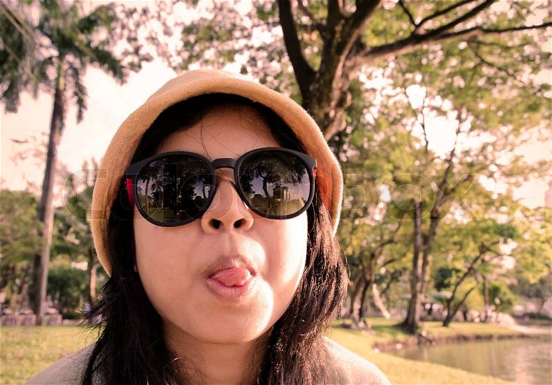 Young naughty woman in yellow cap looking at camera through sunglasses and sticking out tounge, sepia toned, stock photo