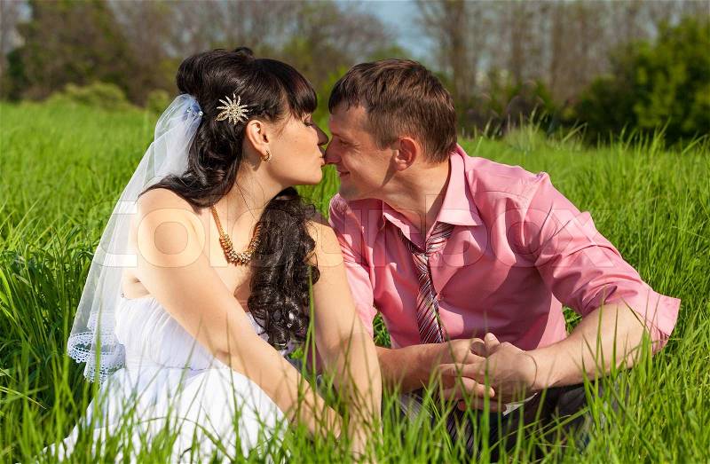 Portrait of bride and groom kissing in lips while sitting on meadow, stock photo