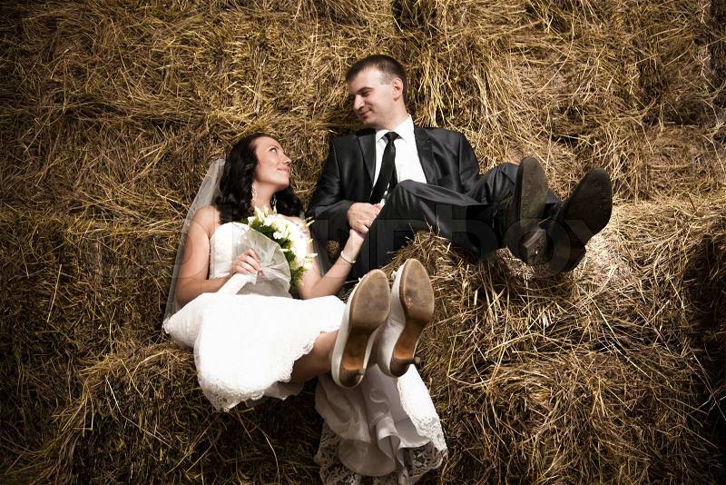 Photo of just married couple holding hands and sitting on hay at stable, stock photo