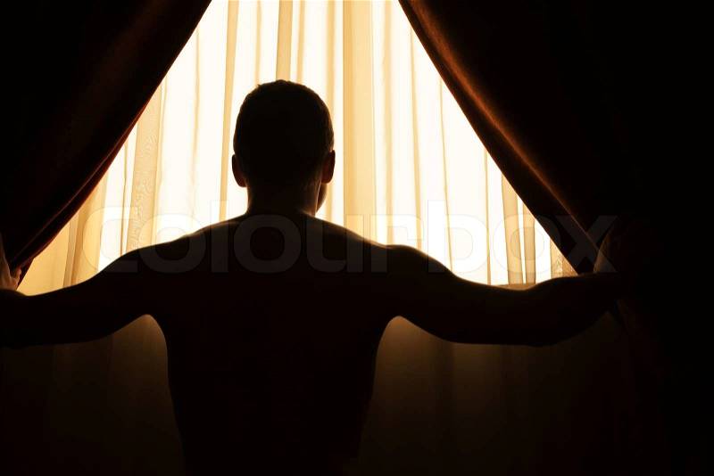 Man in dark room opens curtains on window to the morning sunlight, stock photo