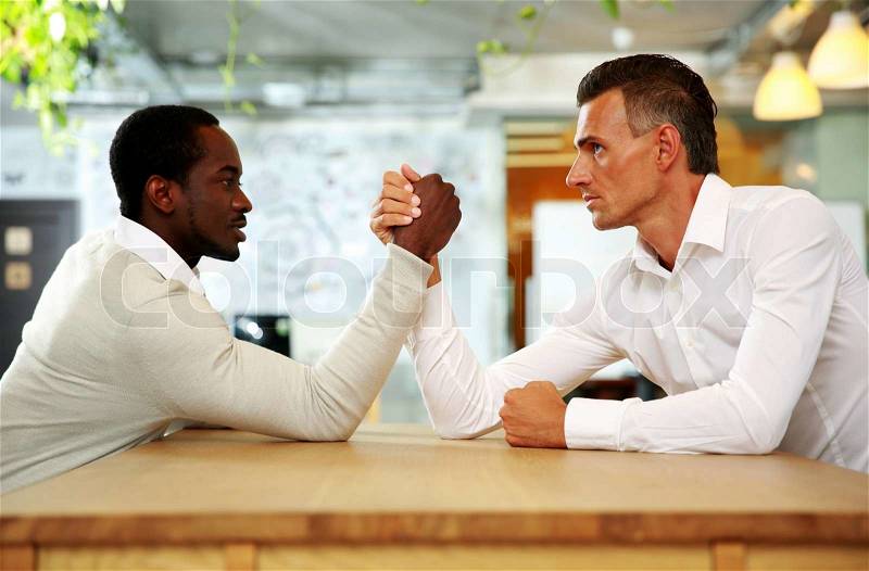 Portrait of two businessmen sitting opposite each other elbowing on the table with their arms grappled in fight, stock photo