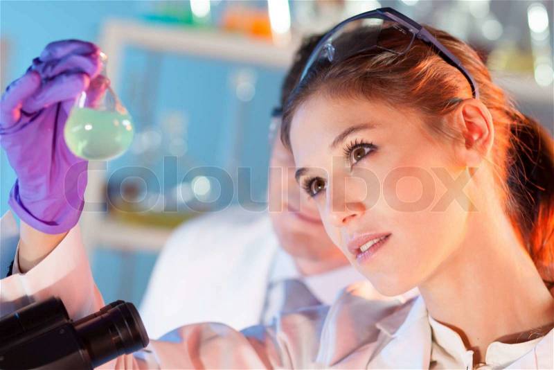 Chemical laboratory scene: attractive young student and her post doctoral supervisor scientist observing the green indikator solution color shift in glass flask, stock photo