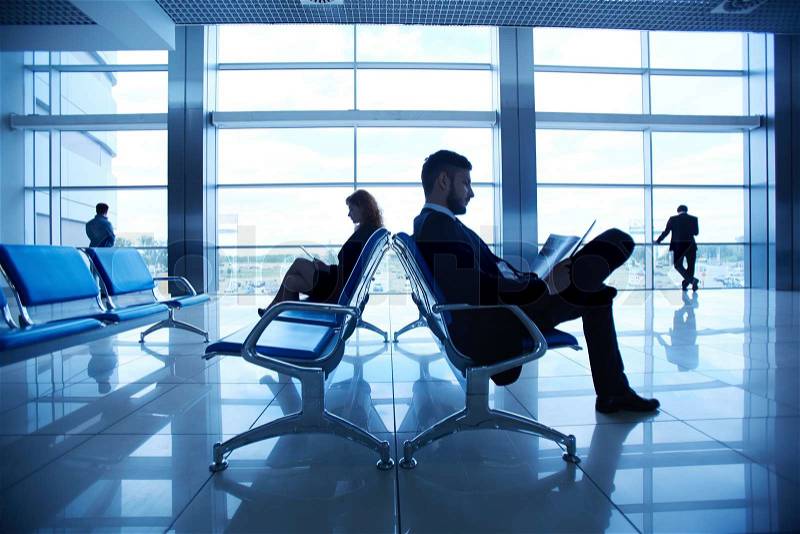 Two business partners reading at the airport on background of their colleagues by the window, stock photo