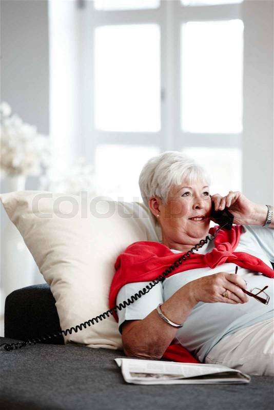 An elderly caucasian woman sitting on the sofa and talking on the phone, stock photo