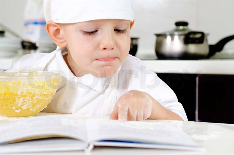 Small boy learning to cook checking his mixture in the mixing bowl against the ingredients in the recipe book, stock photo