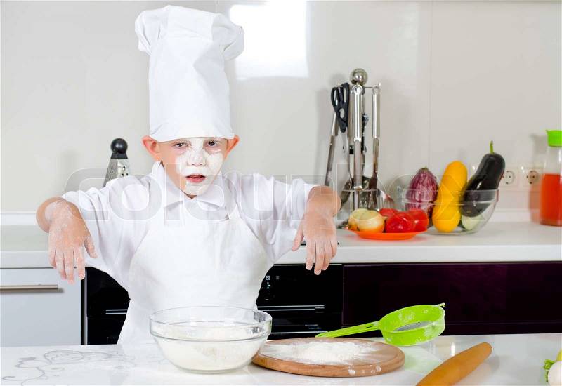 Cute little boy chef in the kitchen with a faceful of flour or batter from the contents of his mixing bowl looking at the camera with a comical expression of disgust, stock photo