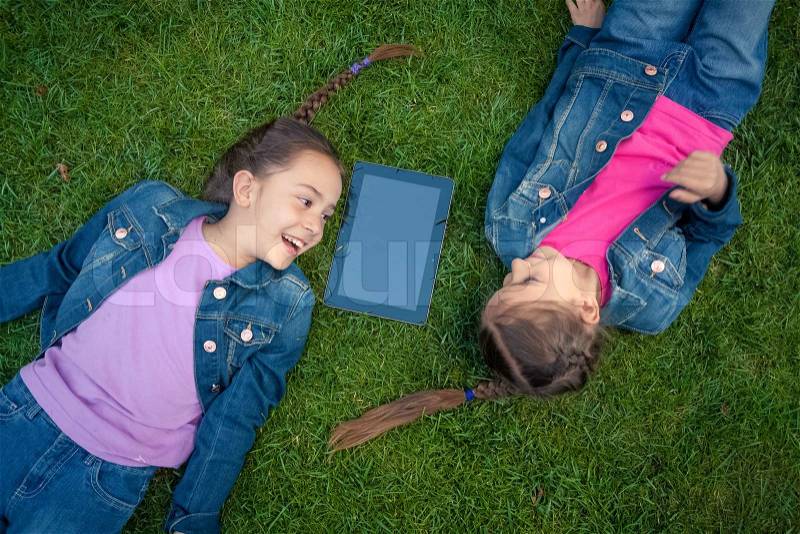 Two little girls lying face to face on grass and looking at tablet, stock photo