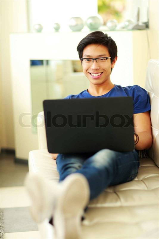 Young happy asian man lying on the sofa with laptop at home, stock photo