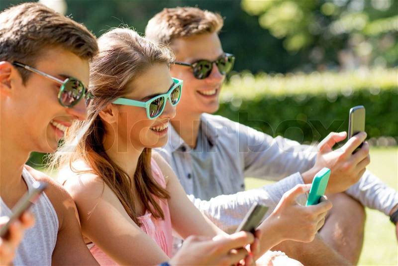 Friendship, leisure, summer, technology and people concept - group of smiling friends with smartphones sitting on grass in park, stock photo