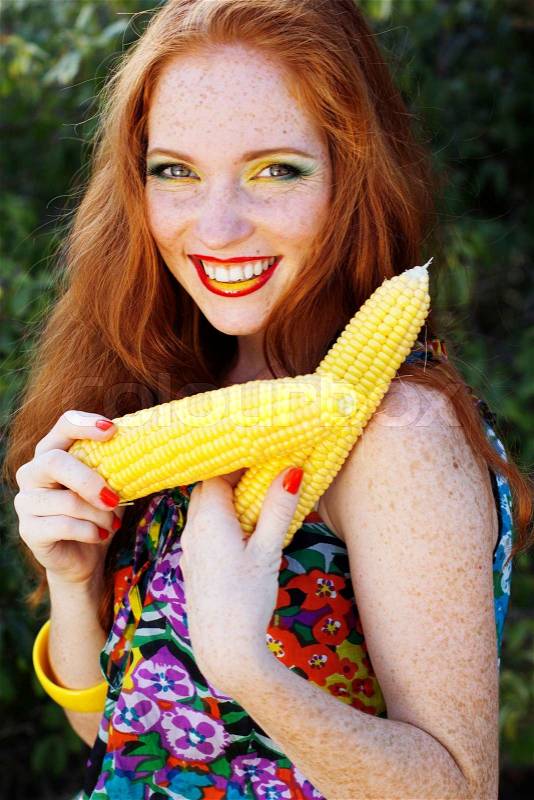 Portrait of a red-heared girl with freckles holding an appetizing corn cob, stock photo