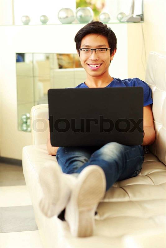 Cheerful asian man lying on the sofa with laptop at home, stock photo