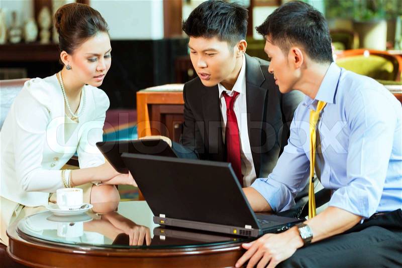 Three Asian Chinese office people or businessmen and businesswoman having a business meeting in a hotel lobby discussing documents on a tablet computer while drinking coffee , stock photo