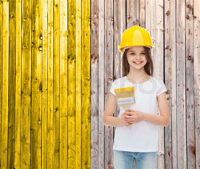 Painting, building, childhood and people concept - smiling little girl in protective helmet with paint brush over wooden fence background, stock photo