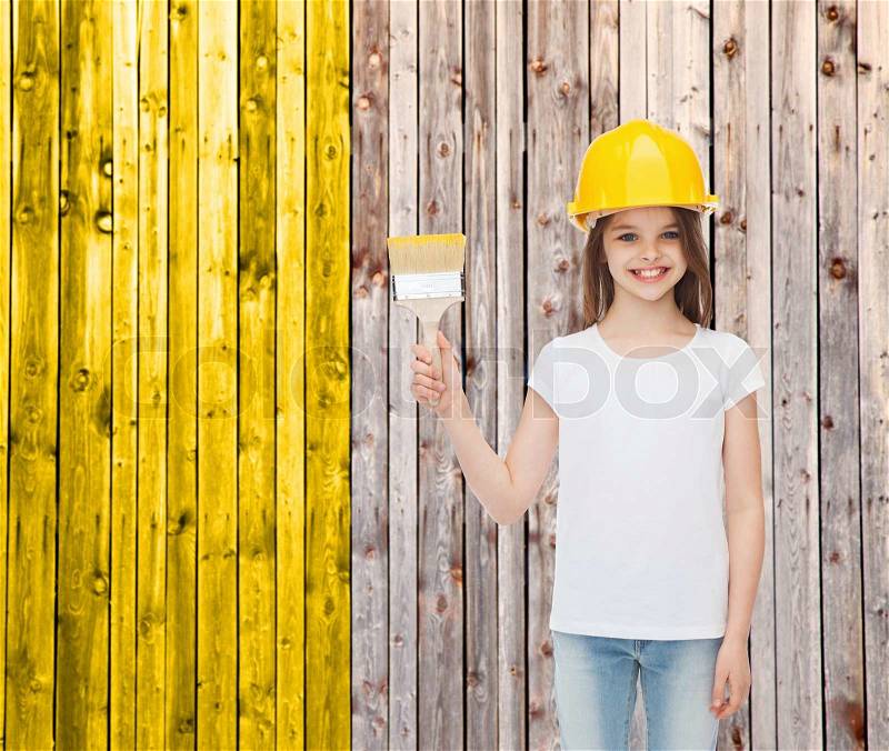 Painting, building, childhood and people concept - smiling little girl in protective helmet with paint brush over wooden fence background, stock photo