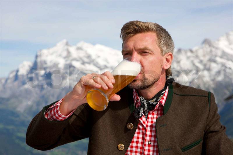 A man sitting at a table with a glass of bear in his hand drinking in front of the mountains and he looks as if he enjoys it, stock photo