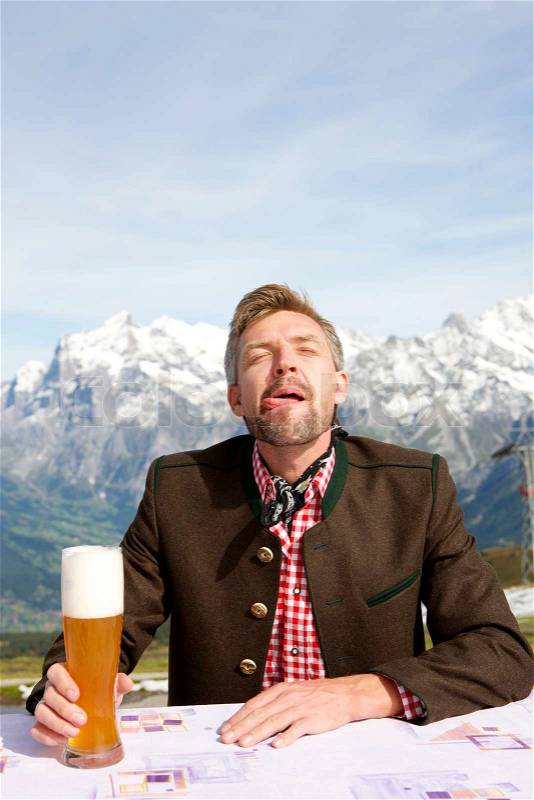A man sitting at a table with a glass of bear in his hand drinking in front of the mountains he is smiling and making a funny face, stock photo