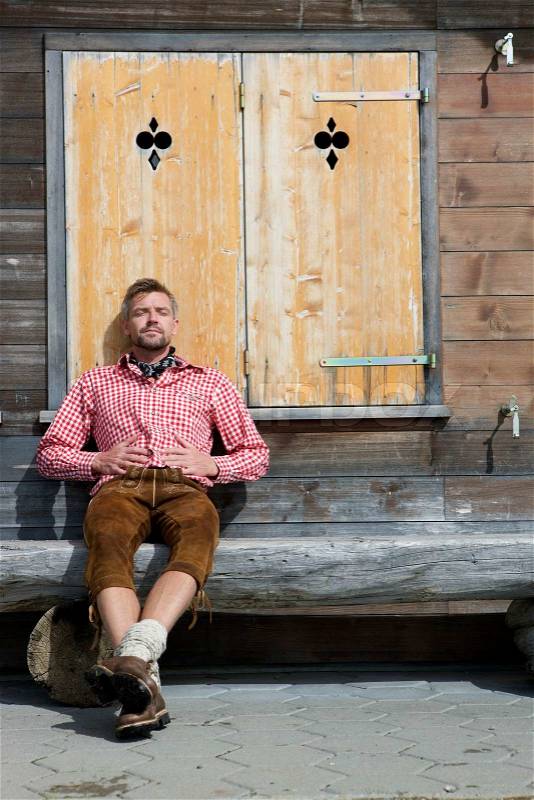 A man resting with closed eyes at a bench in front of a wooden cottage with shuttered windows. He is wearing a red shirt, stock photo
