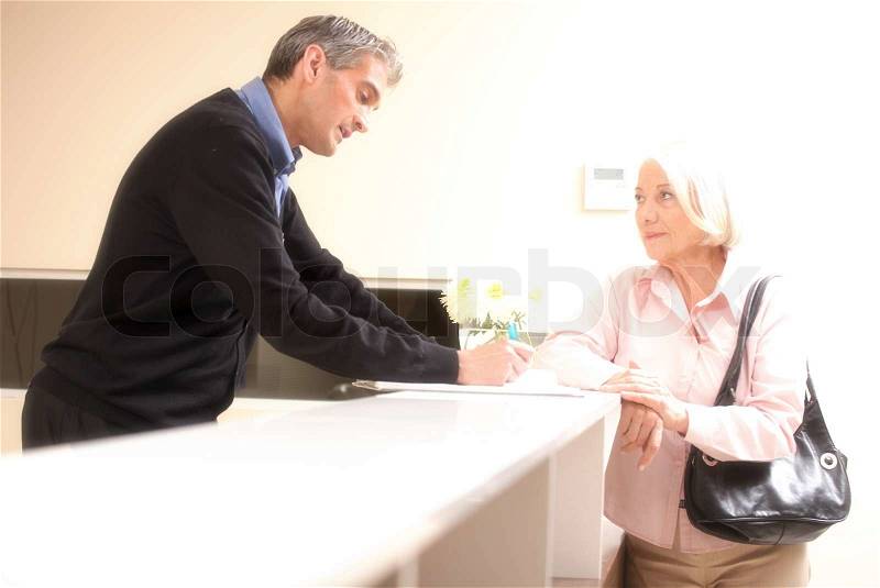 Senior female patient at hospital reception desk with man in 40s collecting personal data, stock photo
