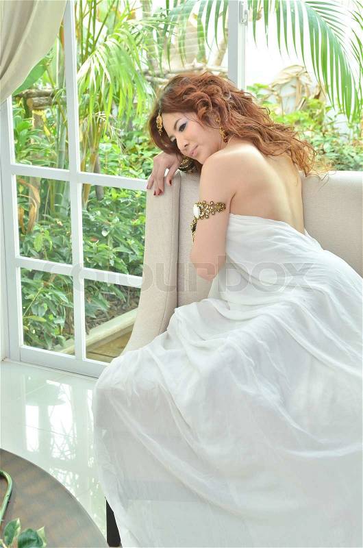 Beautiful young woman in white dress sitting on sofa in her house, stock photo