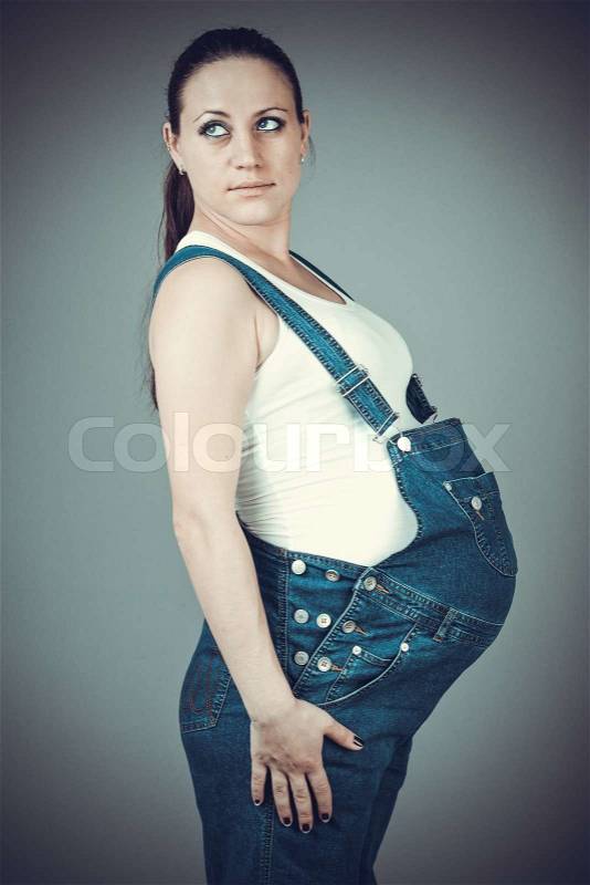 Pregnant beautiful girl in denim overall. Pregnant woman posing on a gray background, stock photo
