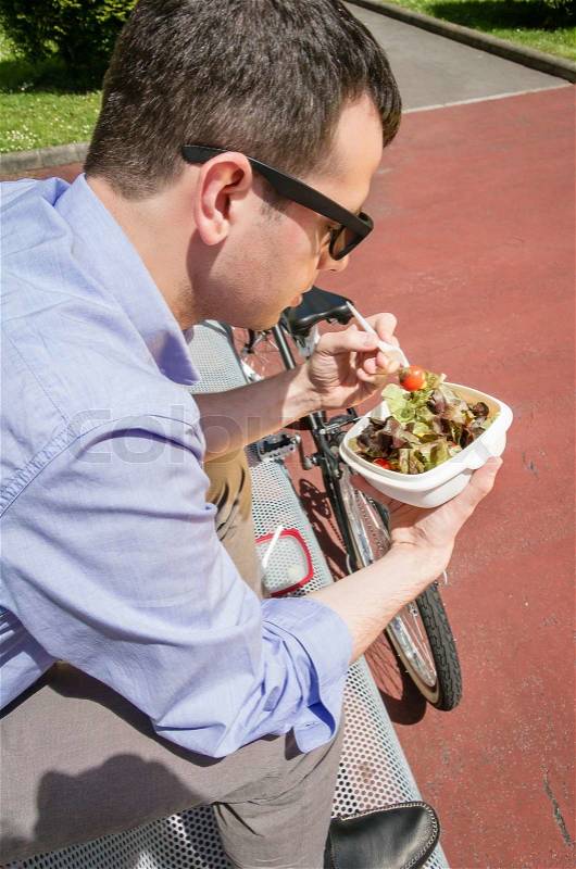 Young business man eating a salad at lunch break sitting on a bench outdoors, stock photo