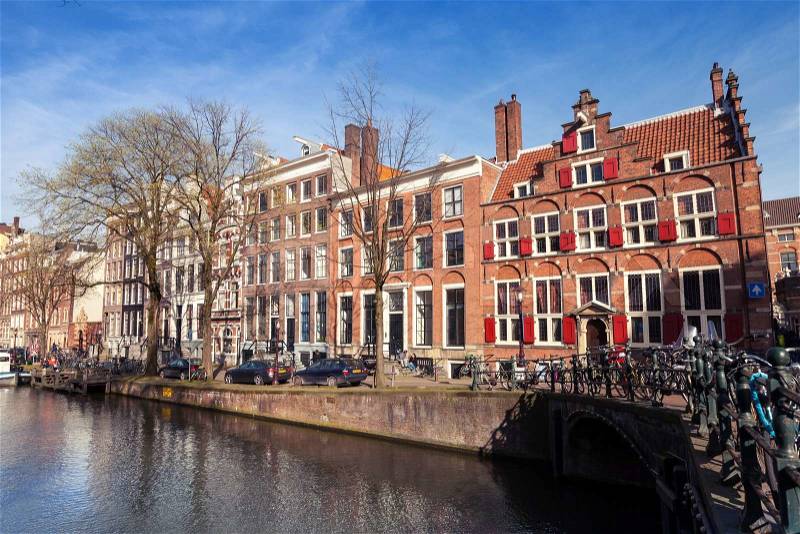 AMSTERDAM, NETHERLANDS - MARCH 19, 2014: Colorful houses along the canal embankment in spring day. Ordinary people walk on the coast, stock photo