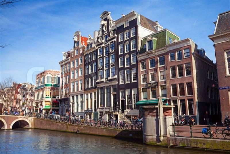 AMSTERDAM, NETHERLANDS - MARCH 19, 2014: Colorful houses on the canal embankment in spring sunny day. Ordinary people walk on the coast, stock photo