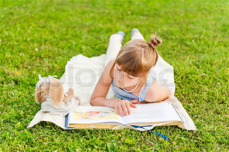 Portrait of adorable little girl resting outdoors and reading a book on a nice summer evening, stock photo