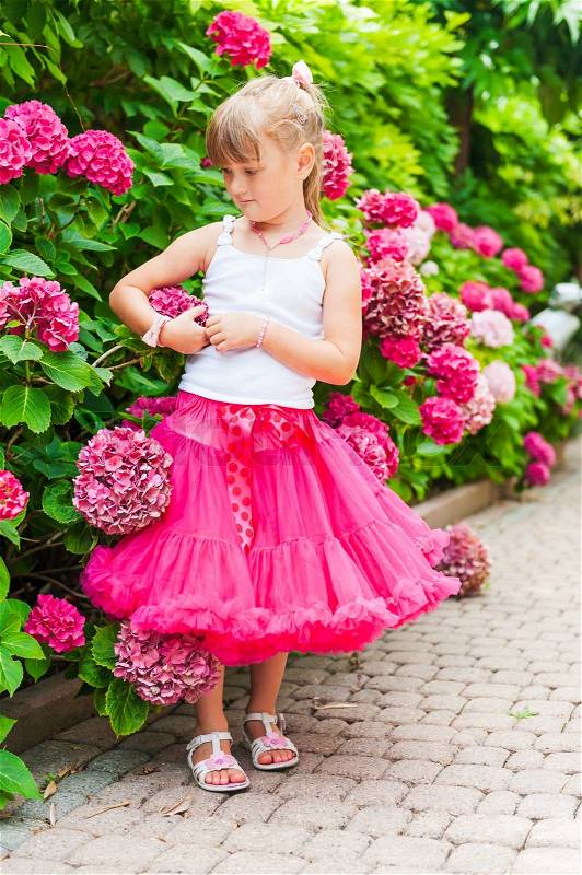 Beautiful little girl wearing bright pink tutu skirt and white top, standing next to bushes of hydrangea, vertical portrait, stock photo