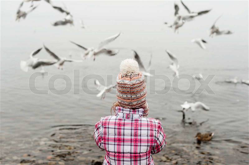 Little girl feeding birds on the lake in winter time, wearing warm jacket and hat, stock photo