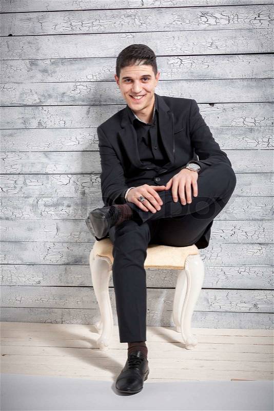 Full length portrait of smiling latin man sitting on chair against wooden wall, stock photo