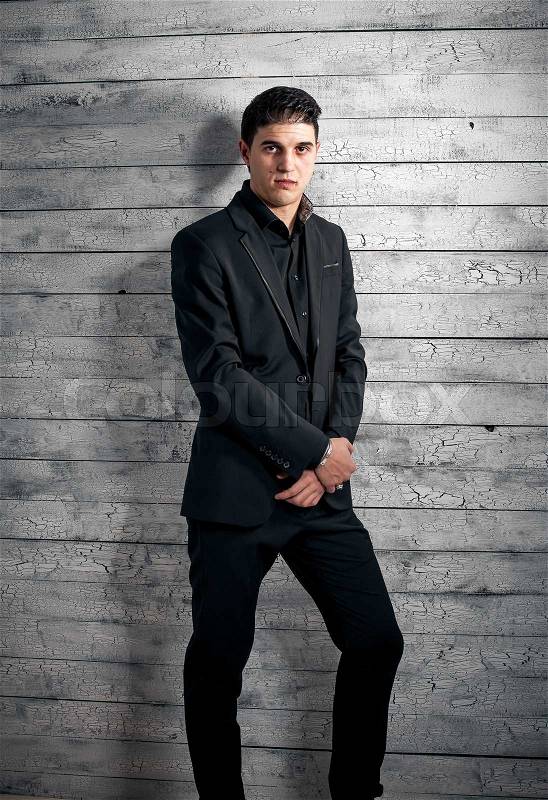 Portrait of brunette man in black suit leaning against white leaning wall, stock photo