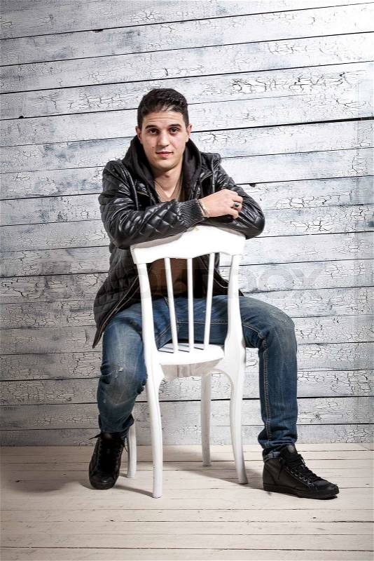Handsome brunette man in jeans and jacket sitting on chair against wooden wall, stock photo