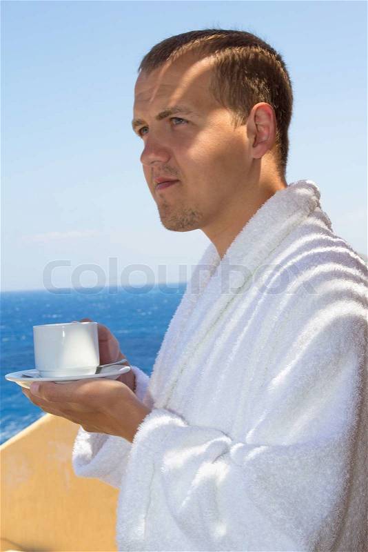 Man in bathrobe with cup of coffee standing at terrace with sea view, stock photo