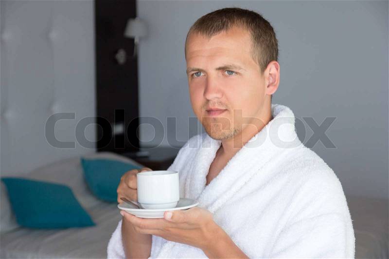Happy young man in bathrobe drinking coffee in hotel room, stock photo