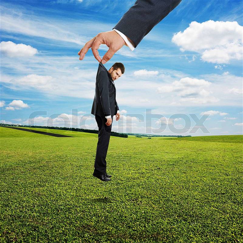 Big hand holding small sad businessman over green field and blue sky, stock photo