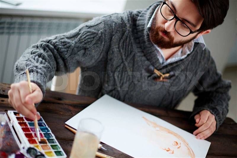 Young man in sweater drawing female portrait with water-colors, stock photo