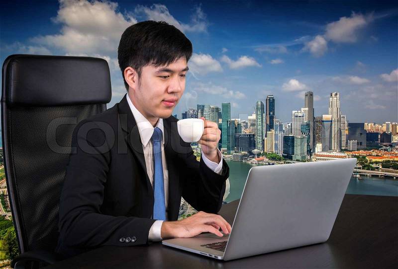 Portrait of goodlooking Asian man sitting at table at working office with laptop computer, smiling at camera. and Singapore city backgroung, stock photo