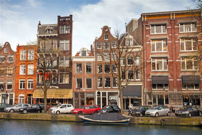 AMSTERDAM, NETHERLANDS - MARCH 19, 2014: Colorful living houses along the canal embankment in spring day. Ordinary people walk on the coast, stock photo