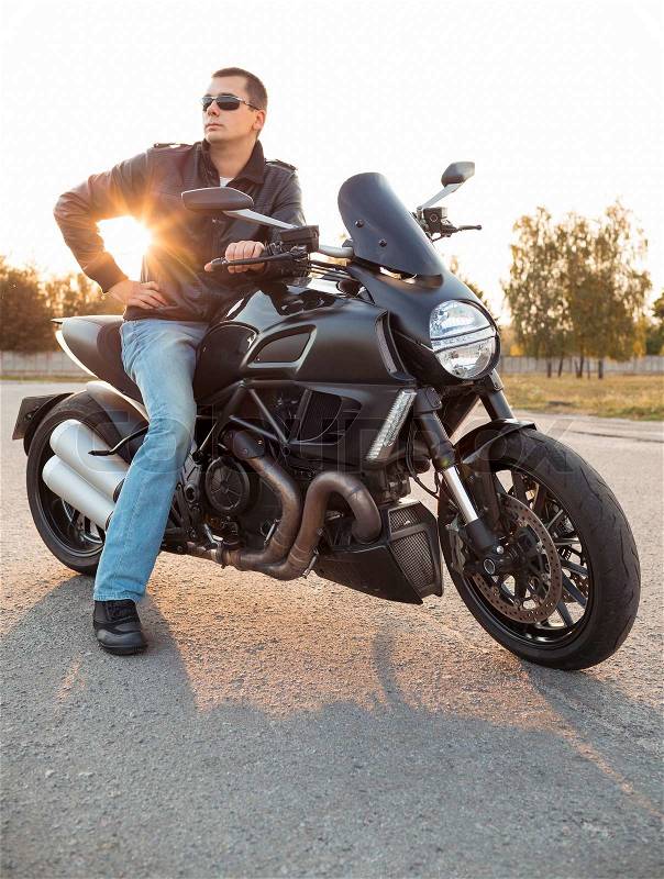 Biker man wearing a leather jacket and sunglasses sitting on his motorcycle outdoors, stock photo