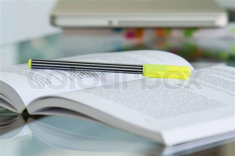 Open book with a yellow pen on top, and a blurry computer in the background, stock photo