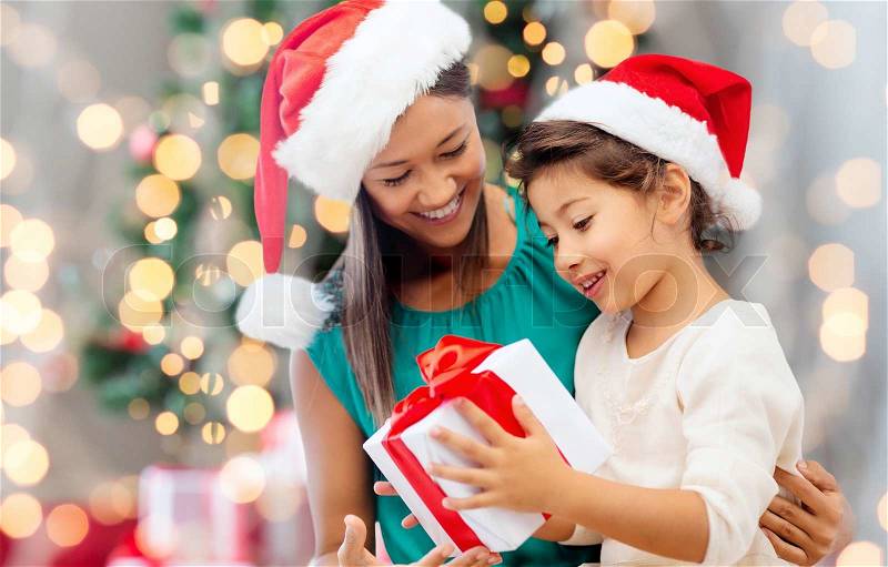 Holidays, celebration, family and people concept - happy mother and little girl in santa helper hats with gift box over living room and christmas tree background, stock photo