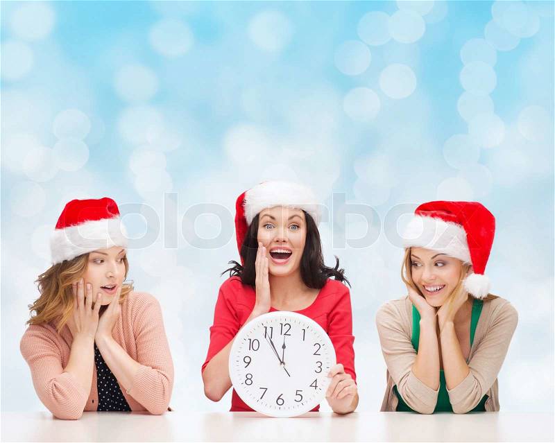 Christmas, winter, holidays, time and people concept - smiling women in santa helper hats with clock over blue lights background, stock photo