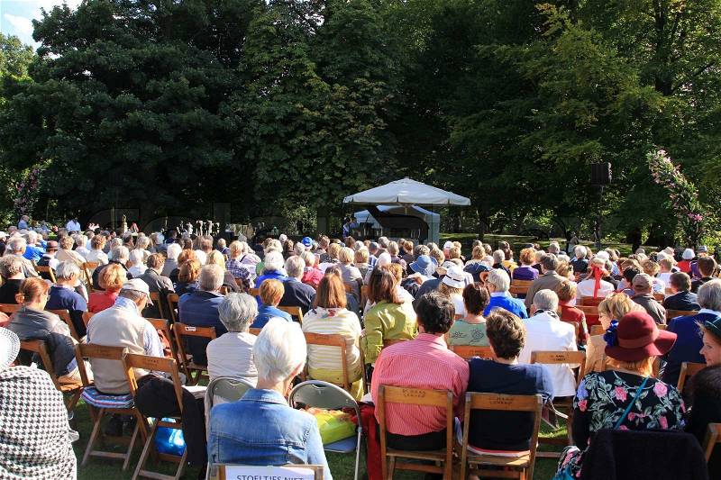Many people sitting on the folding chairs and listen to the concert in the open air in the park in summertime, stock photo