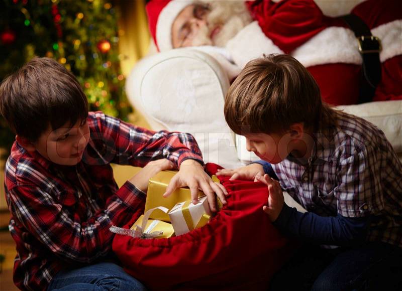 Curious boys choosing gifts from big red sack with Santa Claus sleeping on background, stock photo